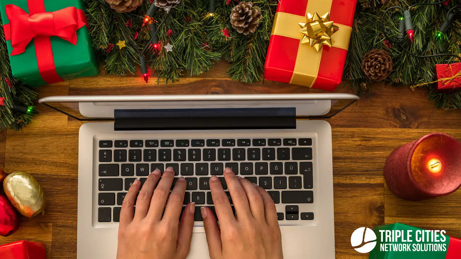 Hands typing on a laptop amid the warmth of Christmas decorations, gifts, and a lit candle—a serene scene perfect for online shopping. In the corner sits a New York IT logo, symbolizing the seamless blend of holiday cheer and cutting-edge technology. Logo in bottom right corner.